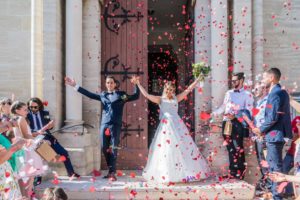 les mariés sortent triomphalement de l'église, levant les bras sous une pluie de confettis rouges en forme de coeur