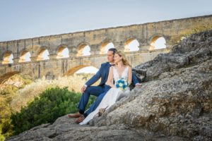 Un couple de mariés assis sur les rochers l'un contre l'autre, devant le pont du gard.