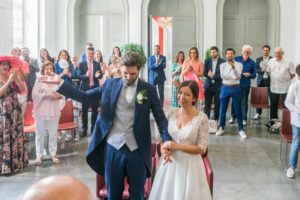 Dans la salle de mariage de la mairie de Nîmes, le marié lève la main, ouverte vers le haut, pour accompagner et augmenter les applaudissements des invités.