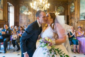 Sous le lustre de la salle de mariage de la mairie de Arles, la mariée donne un baiser à la joue du marié.