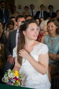 La mariée assise dans la salle de mariage, tenant son bouquet, souriante.