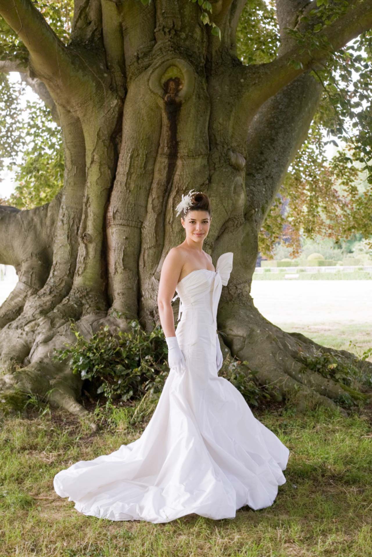 La mariée pause pour le photographe au pied d'un arbre, dans sa robe blanche.