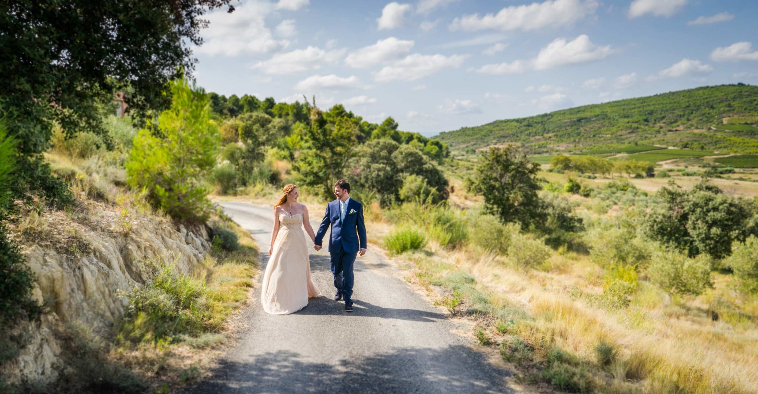 Photographie de Mariage. Les deux mariés marchent sur une route de garrigue main dans la main en se regardant amoureusement. Nîmes, Gard.